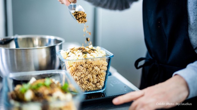 Une personne ajoute des graines (tournesol, courge, etc.) à une portion de pâtes déjà préparée dans un récipient transparent, dans un contexte de cuisine Cookingo.