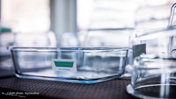 Un récipient rectangulaire en verre transparent avec un autocollant du logo Cookingo est mis en évidence sur une surface sombre, entouré d'autres contenants en verre flous.